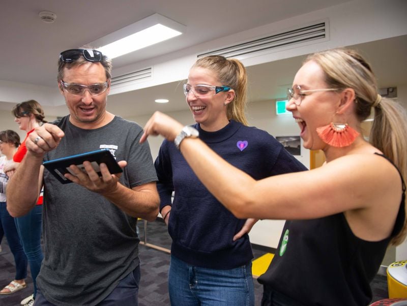 three smiling, laughing teachers wearing safety glasses and pointing to a digital tablet